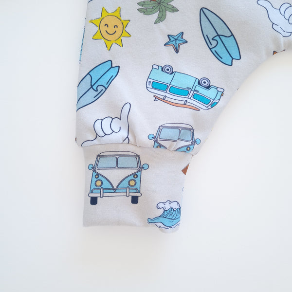 A close-up of a cuff featuring a colorful printed pattern of blue surfboards, a yellow smiling sun, a starfish, a hand gesture, and retro vans, laid flat on a white background.